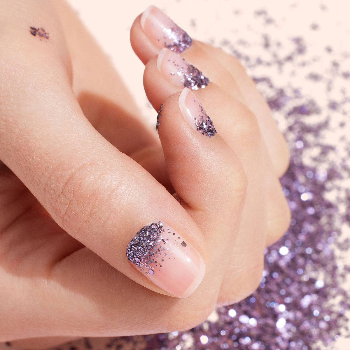 A hand with natural, short nails features a nail art look using Manucurist Lilac biodegradable glitter on the tips. Glitter is scattered on the skin and around the hand, which rests against a light background.