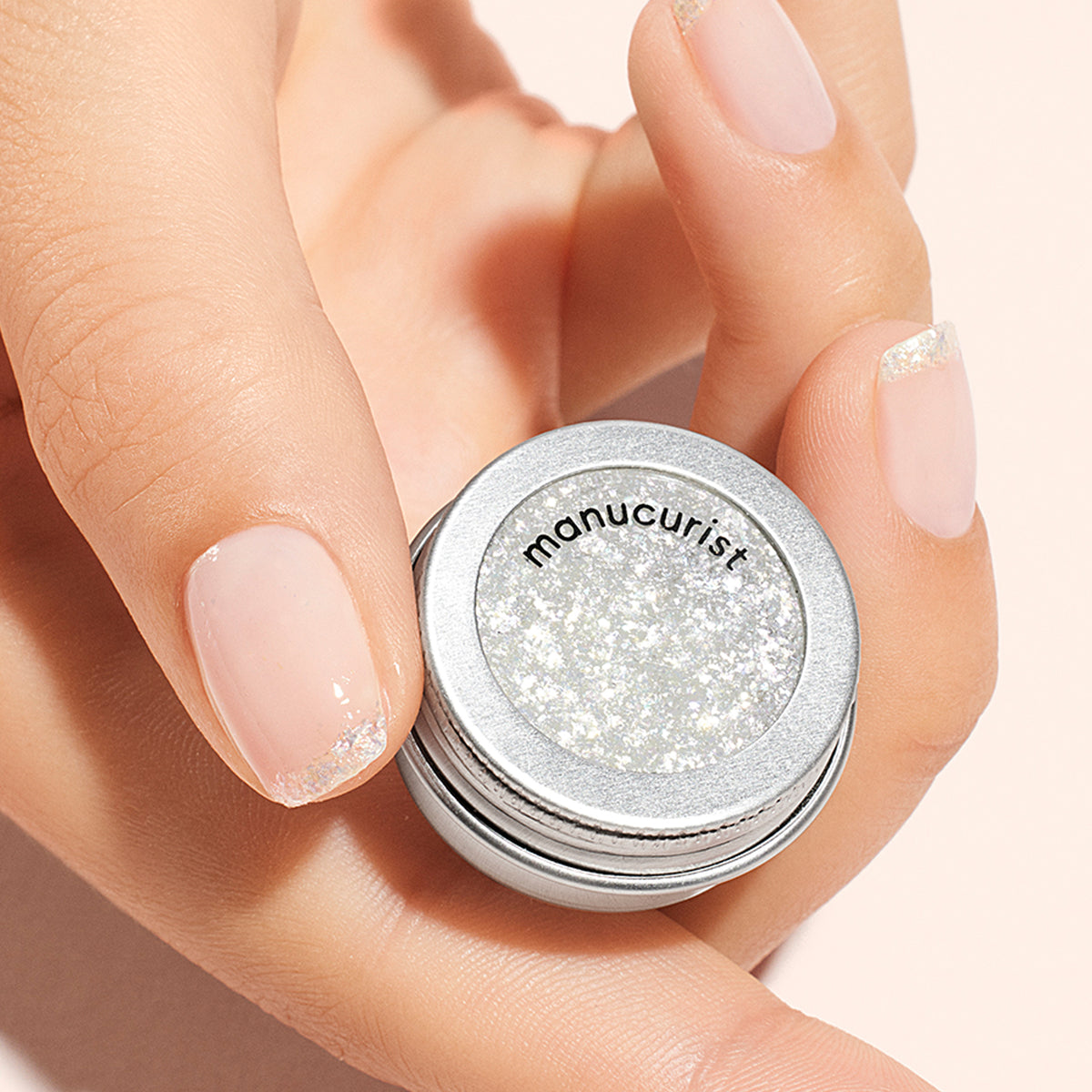 A hand with neatly manicured nails, featuring a subtle glittery French tip and pearlescent flakes, holds a small silver Manucurist container of Nacres Flakes filled with sparkly glitter powder.