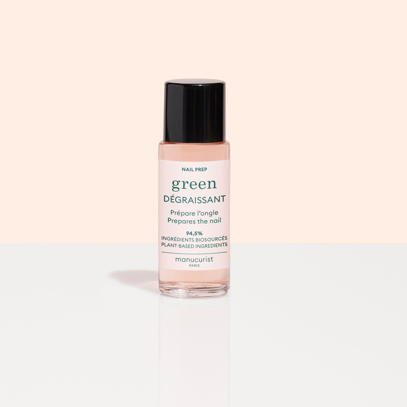 A clear bottle of Manucurist Degreaser with pink liquid and a black cap stands upright on a glossy white surface against a pale peach background, perfect for boosting nail polish adhesion.
