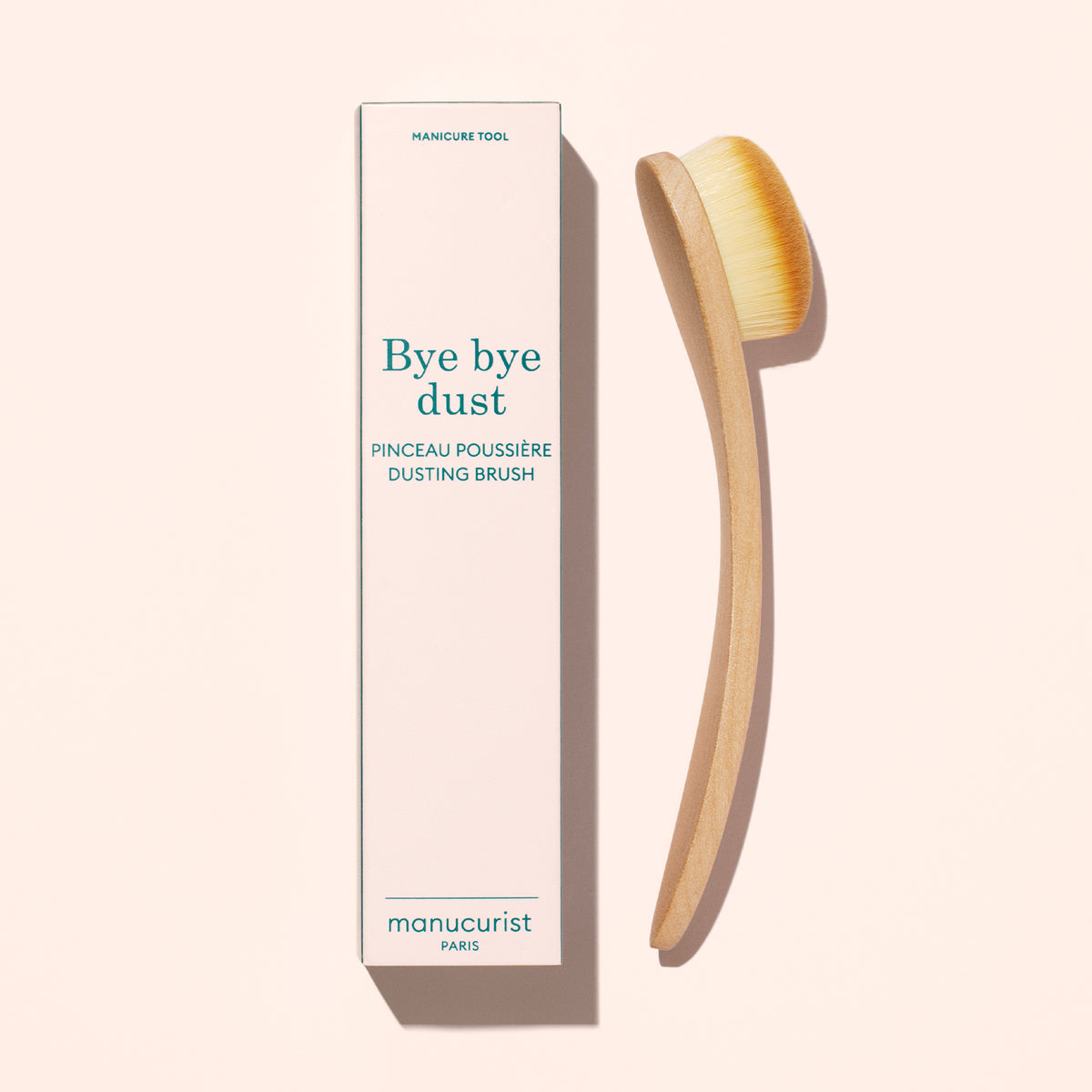 A Manucurist Dust Brush with a wooden handle and dense vegan bristles sits next to a rectangular box labeled "Bye bye dust, Dust Brush." Both are set against a light beige background.