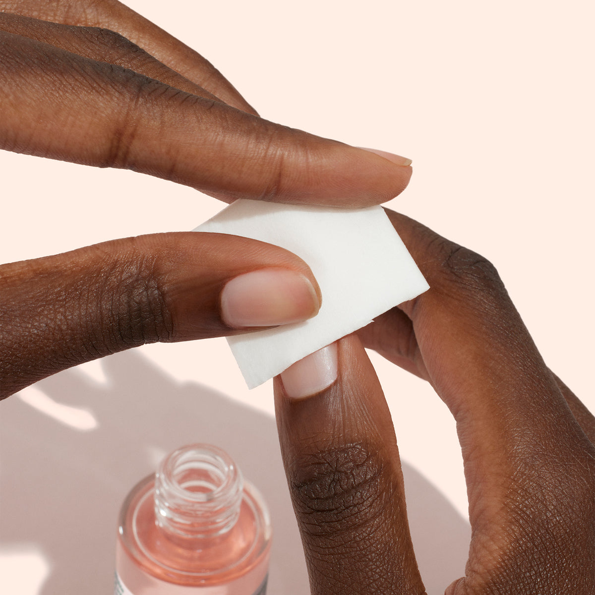 A person uses a white cotton pad with Manucurist Degreaser to clean their nails for proper prep, while an open bottle of the pink liquid sits nearby on a light background.
