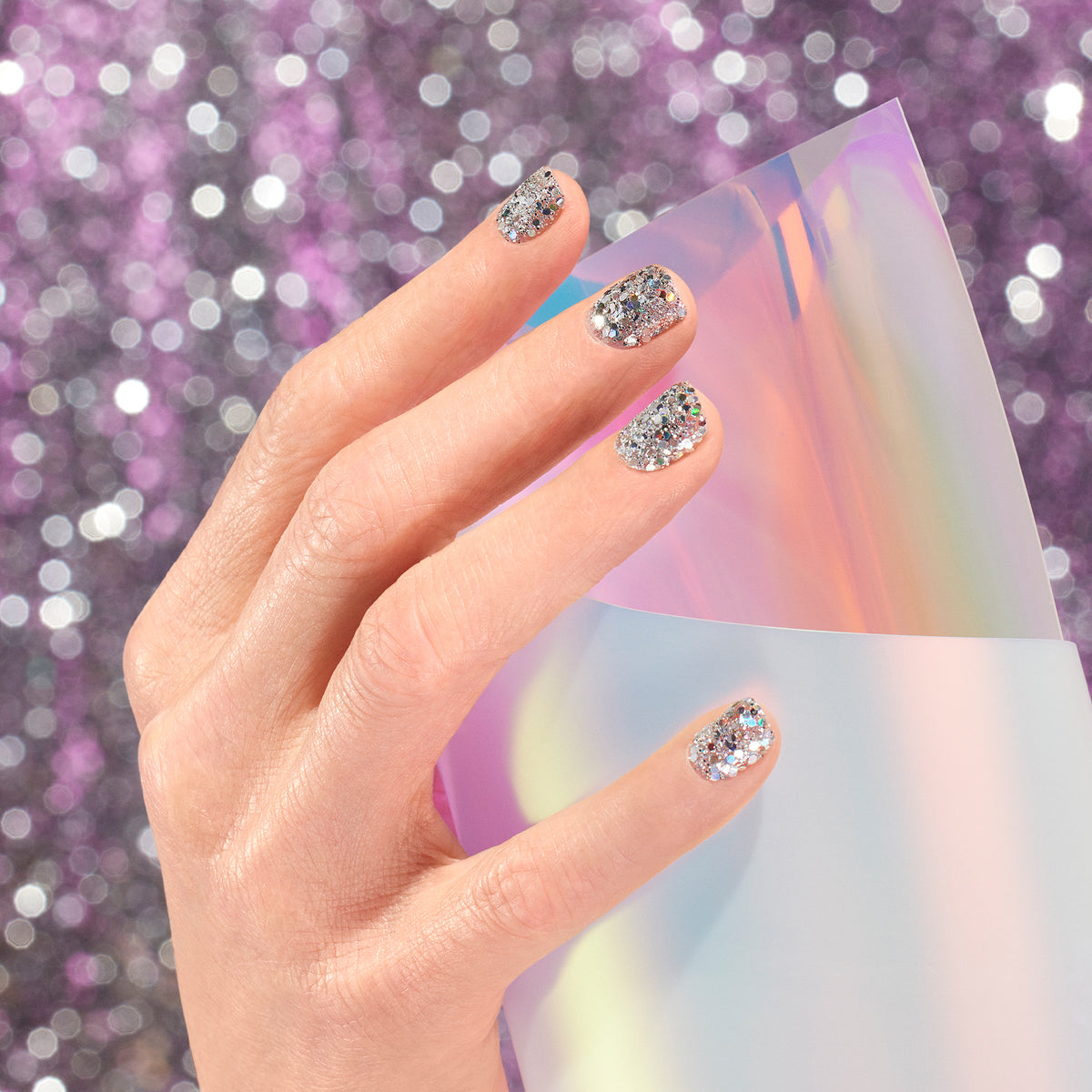 A hand with Manucurist Holo biodegradable glitter polish holds iridescent translucent paper, while a sparkly purple background creates a festive and glamorous vibe.