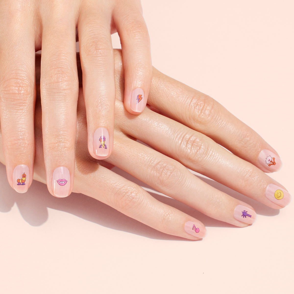 Two hands with natural nails, each manicure decorated with Manucurist Nail Art Stickers—smiley faces, lips, lightning bolts, and other playful designs—set against a soft pink background.