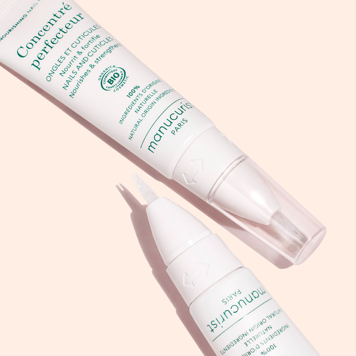 Two Nourishing Nail Pens - Cosmos Organic by Manucurist rest on a pale pink background, one open to show the brush tip—perfect for cuticle care and nail strengthening with this organic routine.