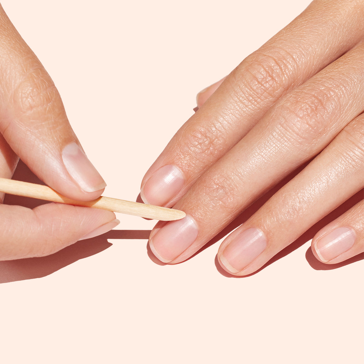 A hand uses the Manucurist UK Wooden cuticle stick x2 to gently push back cuticles on neatly manicured nails against a light beige background.