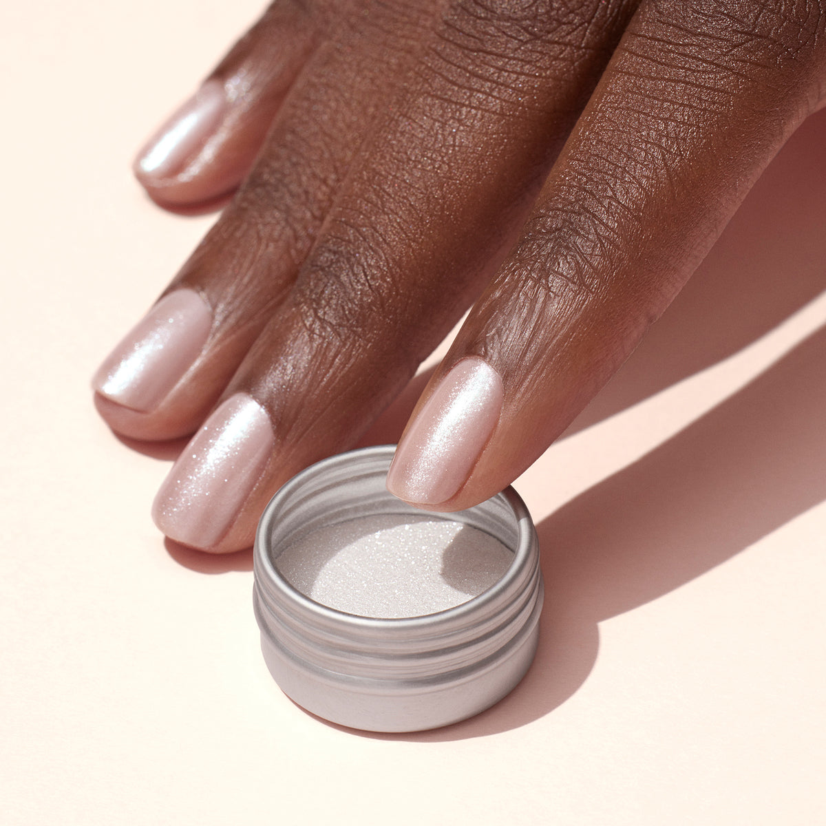 A hand with light pink, shimmery nails featuring a subtle white glazed effect rests beside an open small silver container of Manucurist&