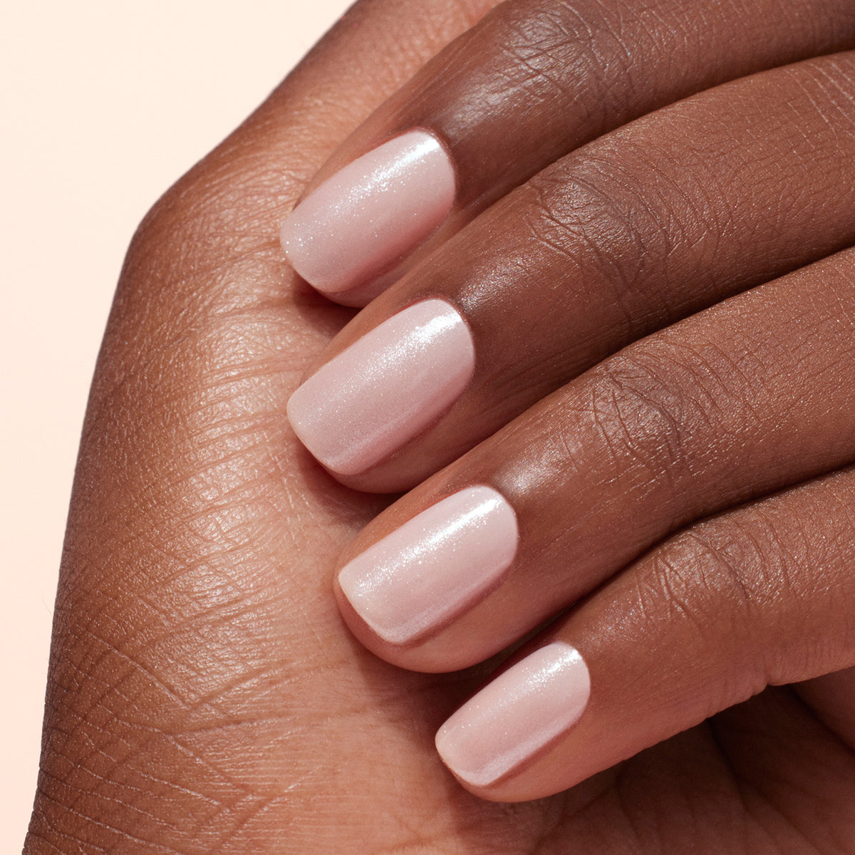 A hand with medium-dark skin tone shows manicured nails painted light pink and topped with Manucurist’s White Glazed Effect Powder, creating a modern, subtle shimmer. The fingers are gently resting against each other.