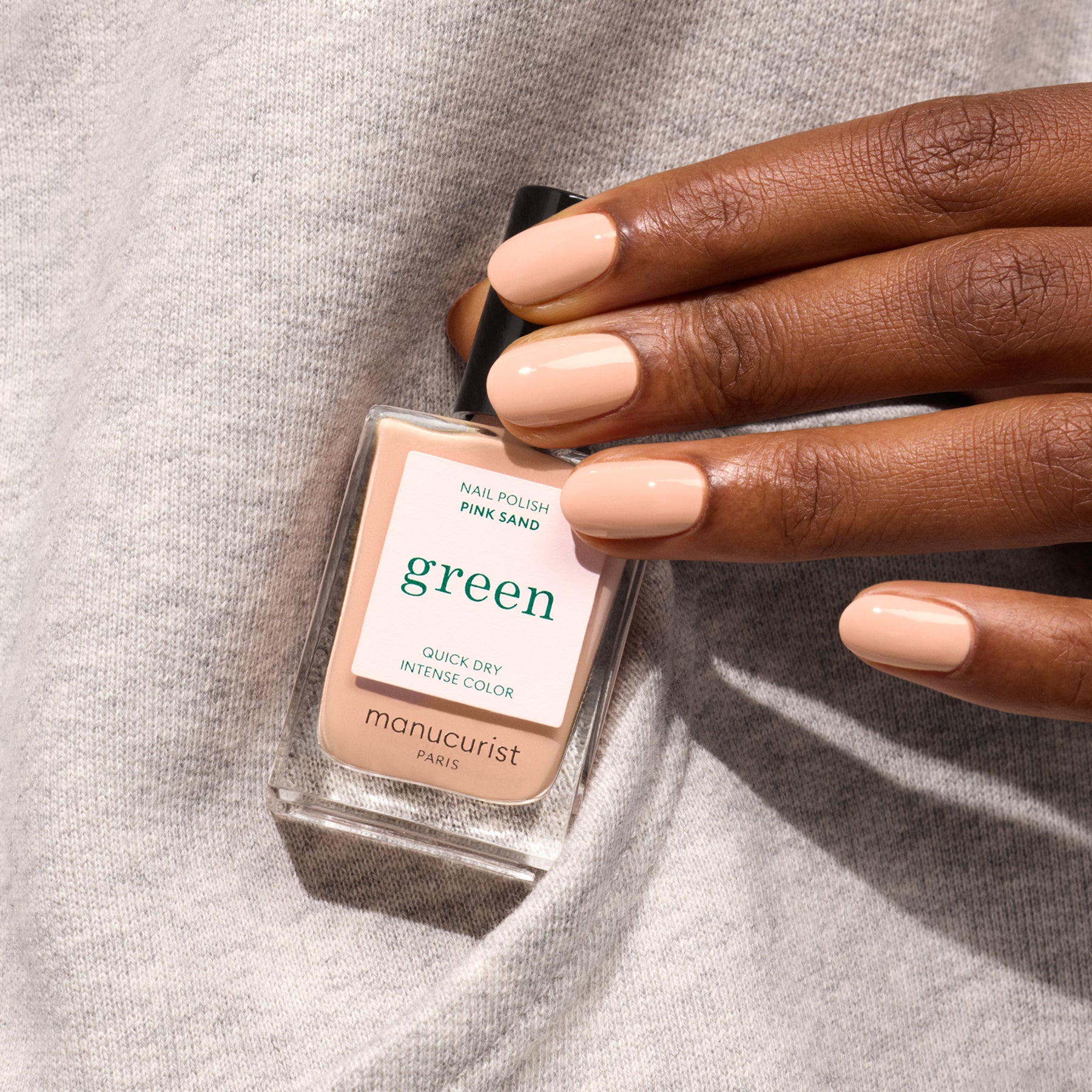 A hand with neatly manicured nails painted in Pink Sand by Manucurist holds the bottle against a light gray fabric background; the polish is a light nude shade with orange undertones.