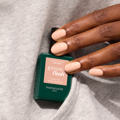 A hand with neatly manicured, nude nails holds a Manucurist Pink Sand gel polish bottle, resting on a gray fabric background.