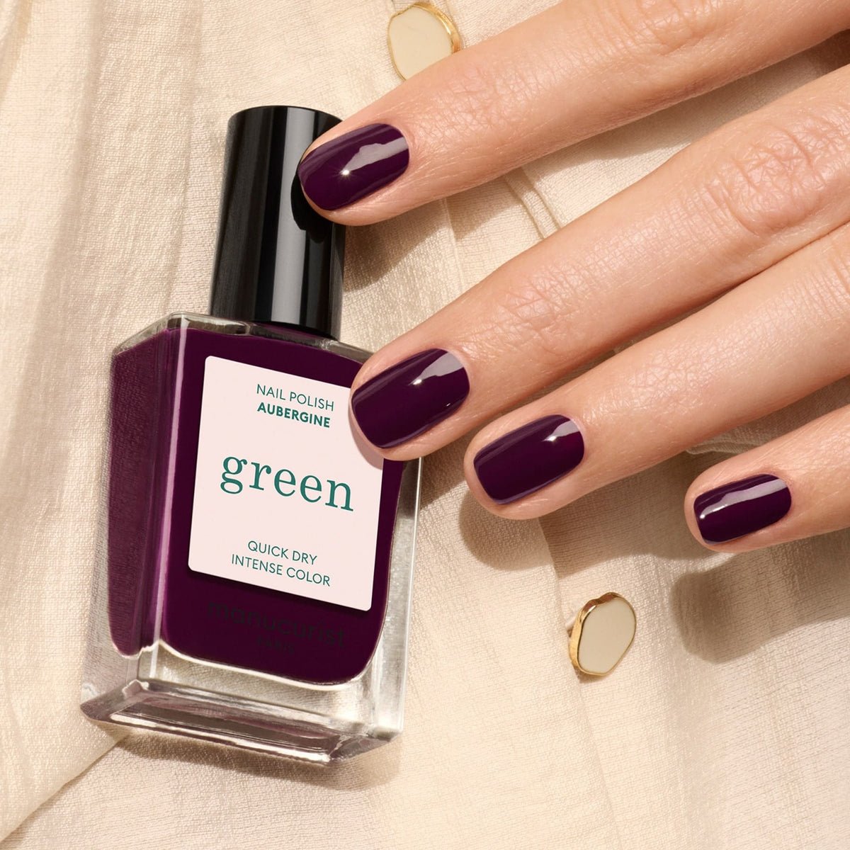 A hand with neatly manicured nails painted deep purple holds a bottle of Manucurist Aubergine nail polish, resting on cream-colored fabric with gold buttons.