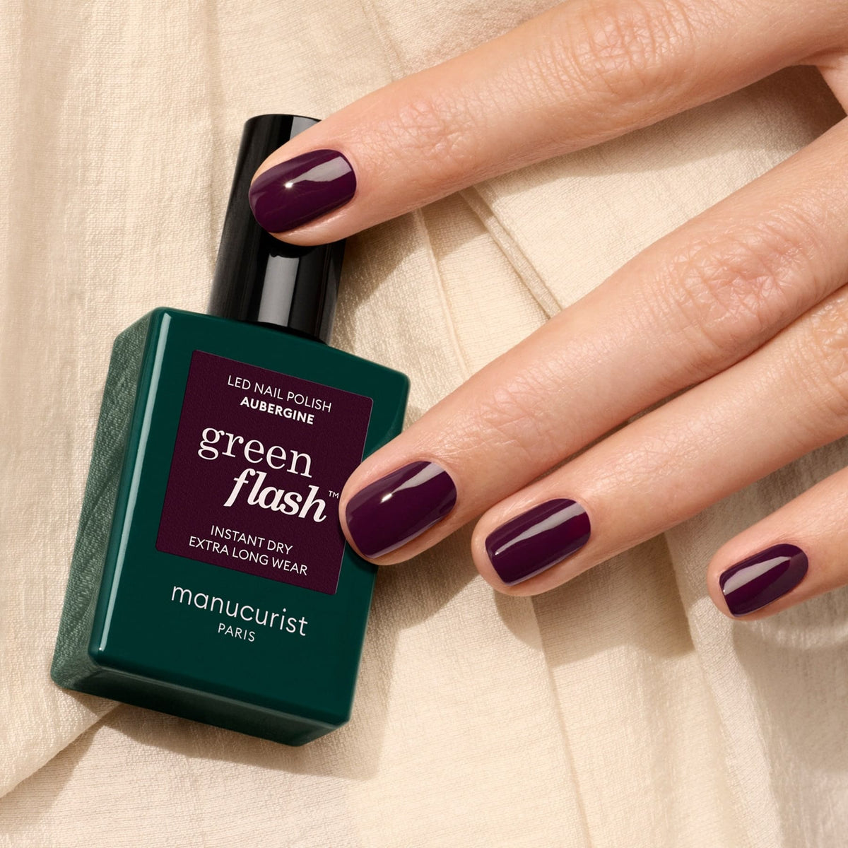 A hand with neatly manicured nails painted deep plum holds a bottle of Manucurist Aubergine polish, resting on beige fabric.