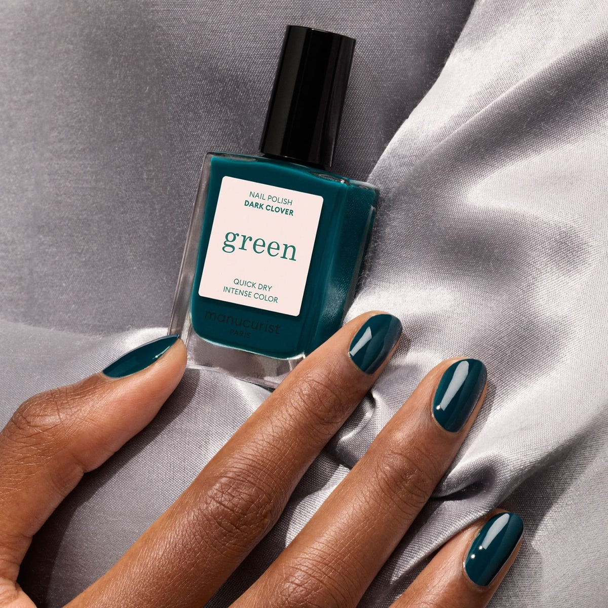 A hand with neatly manicured nails painted in Manucurist’s Dark Clover rests on gray satin, beside a matching bottle—ideal for anyone who loves classic style or wants to try a bold dark green nail polish.