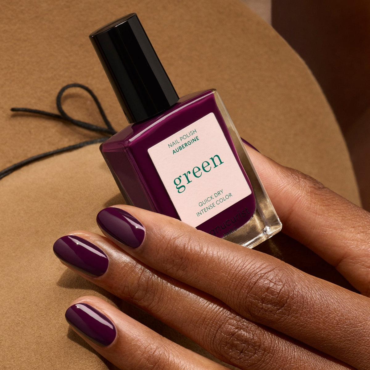 A hand with neatly manicured nails painted in deep purple holds a matching bottle of Manucurist’s Aubergine nail polish, set against a soft brown background.