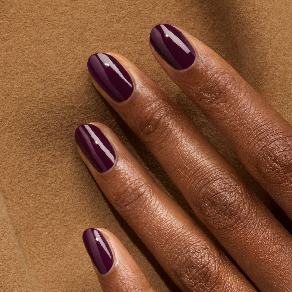 A hand with medium-dark skin tone displays neatly manicured nails painted in Manucurist’s Aubergine, a glossy dark purple gel polish, resting on soft beige fabric.