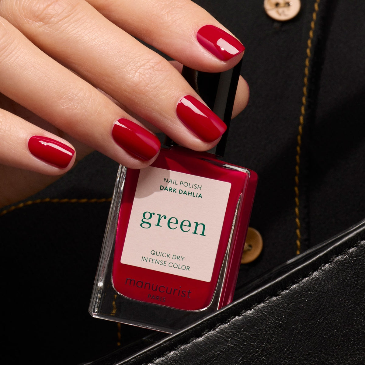 A hand with neatly manicured red nails holds a bottle of Manucurist Dark Dahlia nail polish against a black background with gold button details.