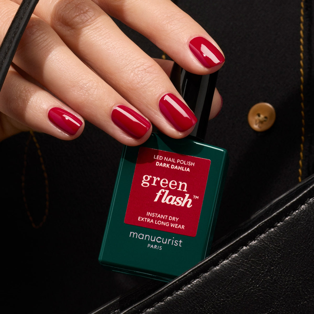 A hand with deep red gel nails holds a Manucurist Dark Dahlia polish bottle against a black background with a gold button.
