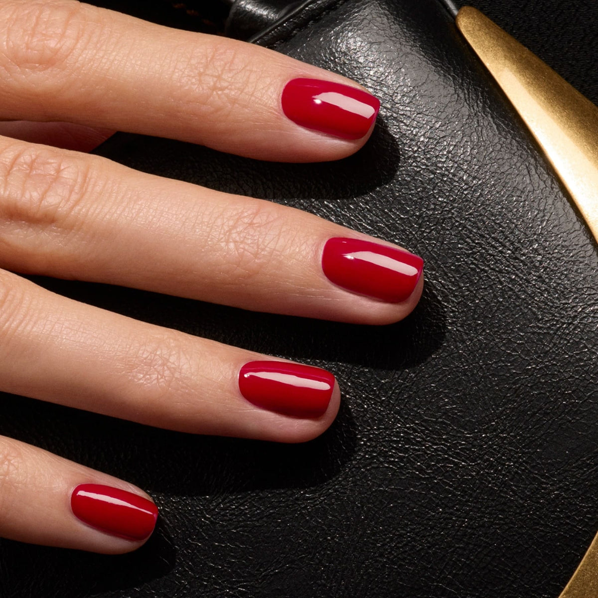 A close-up of a hand with short, neatly manicured nails painted in Dark Dahlia, a dark red polish from Manucurist, resting on black leather with a gold accent.
