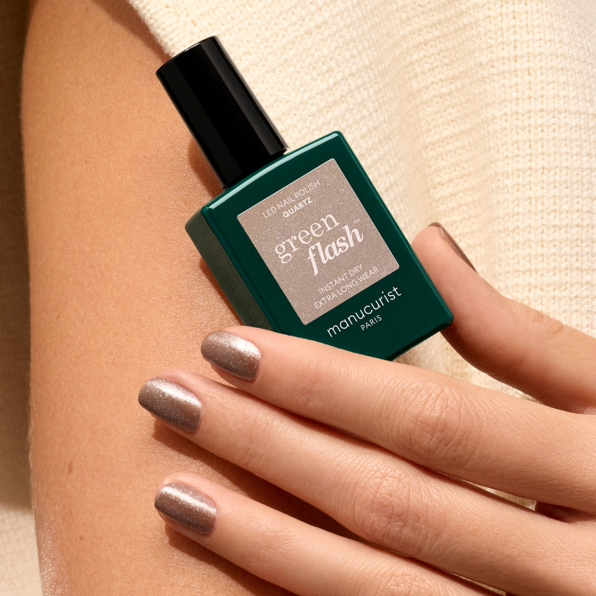 A hand with shimmery nude and chestnut brown nails holds a green bottle of Manucurist Quartz nail polish against light beige textured fabric.