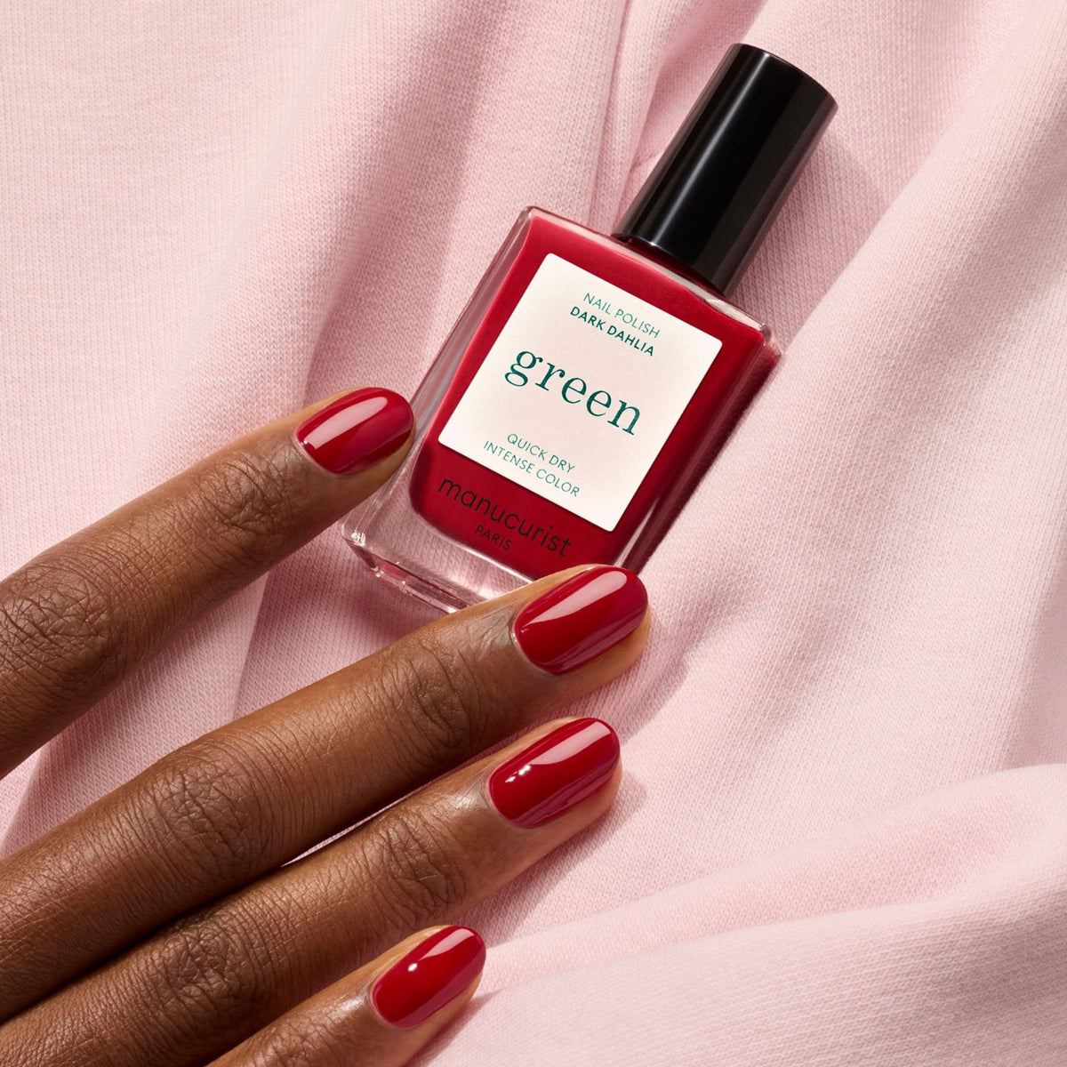 A hand with neatly manicured red nails rests on pink fabric beside a bottle of Manucurist&