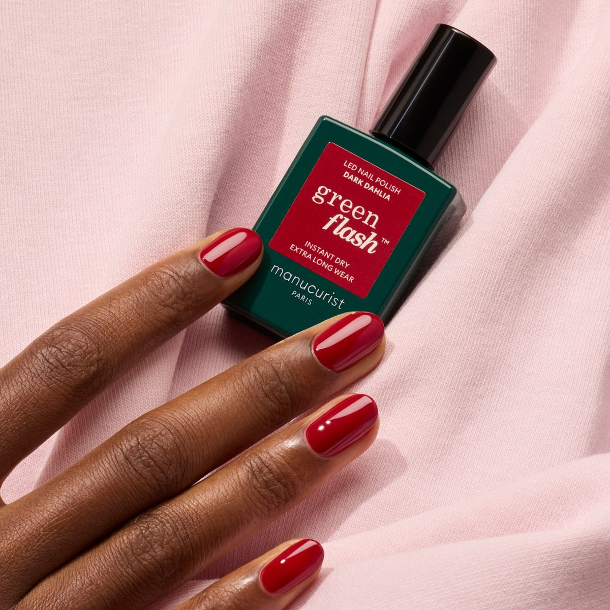 A hand with a glossy deep red gel polish manicure rests on pale pink fabric next to a bottle of Manucurist Dark Dahlia nail polish.