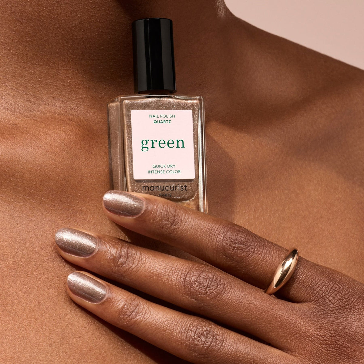 A hand with metallic, silver-beige "Quartz" nail polish by Manucurist holds the bottle against bare skin. A simple gold ring on the middle finger complements the subtle shimmer of this silver glitter polish.