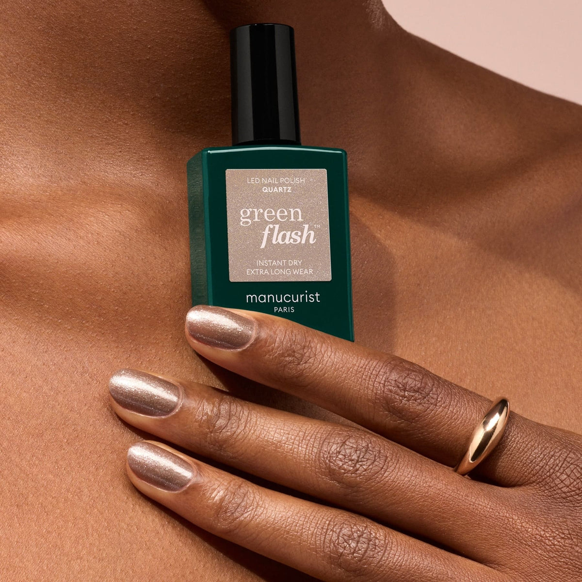 A hand with metallic nude Quartz polished nails holds a green bottle of Manucurist nail polish. The person wears a simple gold ring on their middle finger.