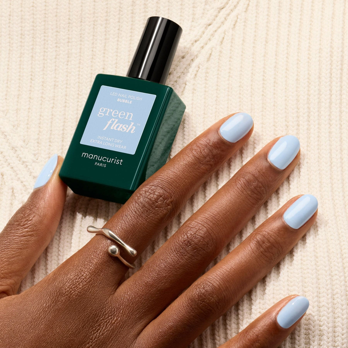 A hand with cool light blue nails rests on cream knit fabric, wearing a silver ring and holding a bottle of Bubble nail polish by Manucurist between the fingers.