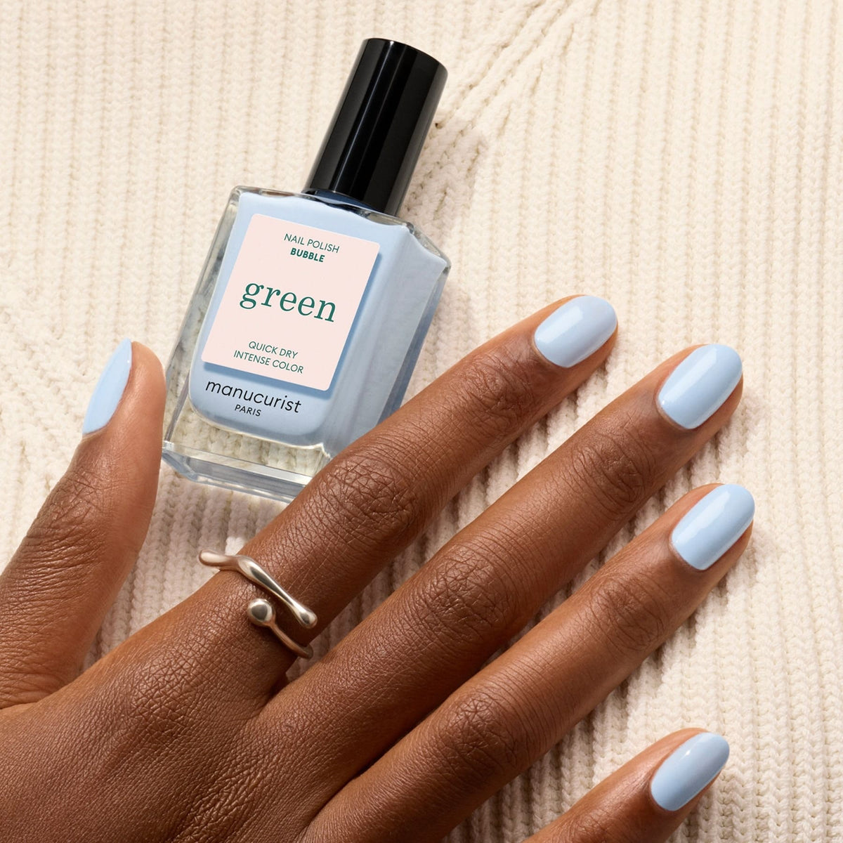 A hand with light blue-grey "Bubble" nail polish by Manucurist rests on cream knit fabric. The person wears a silver ring, and a matching bottle of Manucurist Bubble polish is placed nearby.
