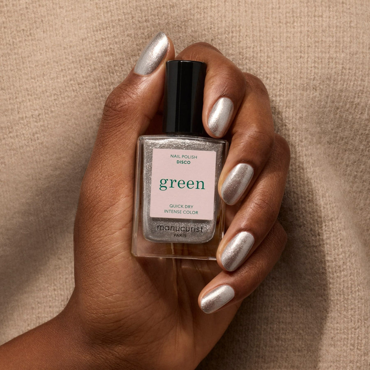 A hand with opaque glittery silver nail polish holds a bottle labeled Disco by Manucurist against a beige textured background.