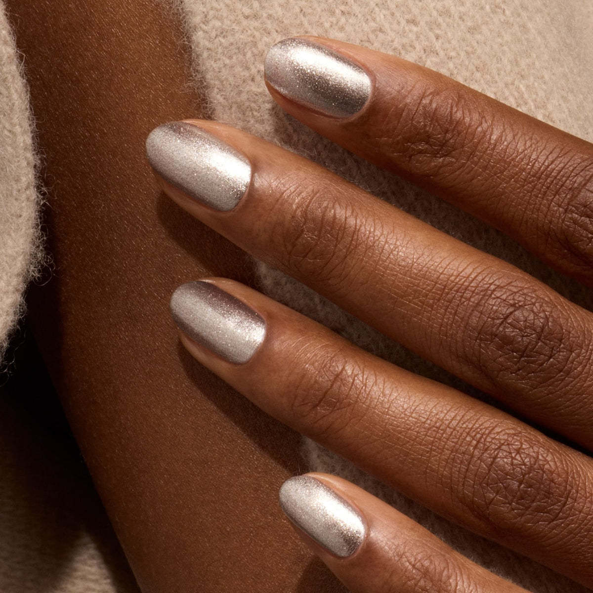 A close-up of a hand with medium-brown skin and short, oval nails painted in Manucurist’s Disco, an opaque glittery silver polish, resting on beige textured fabric.