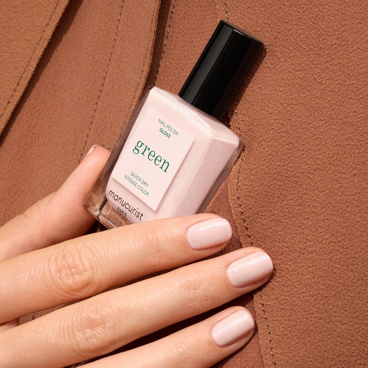 A hand with neatly manicured, pale pink nails holds a bottle of Manucurist Gloss nail polish against a brown textured background.