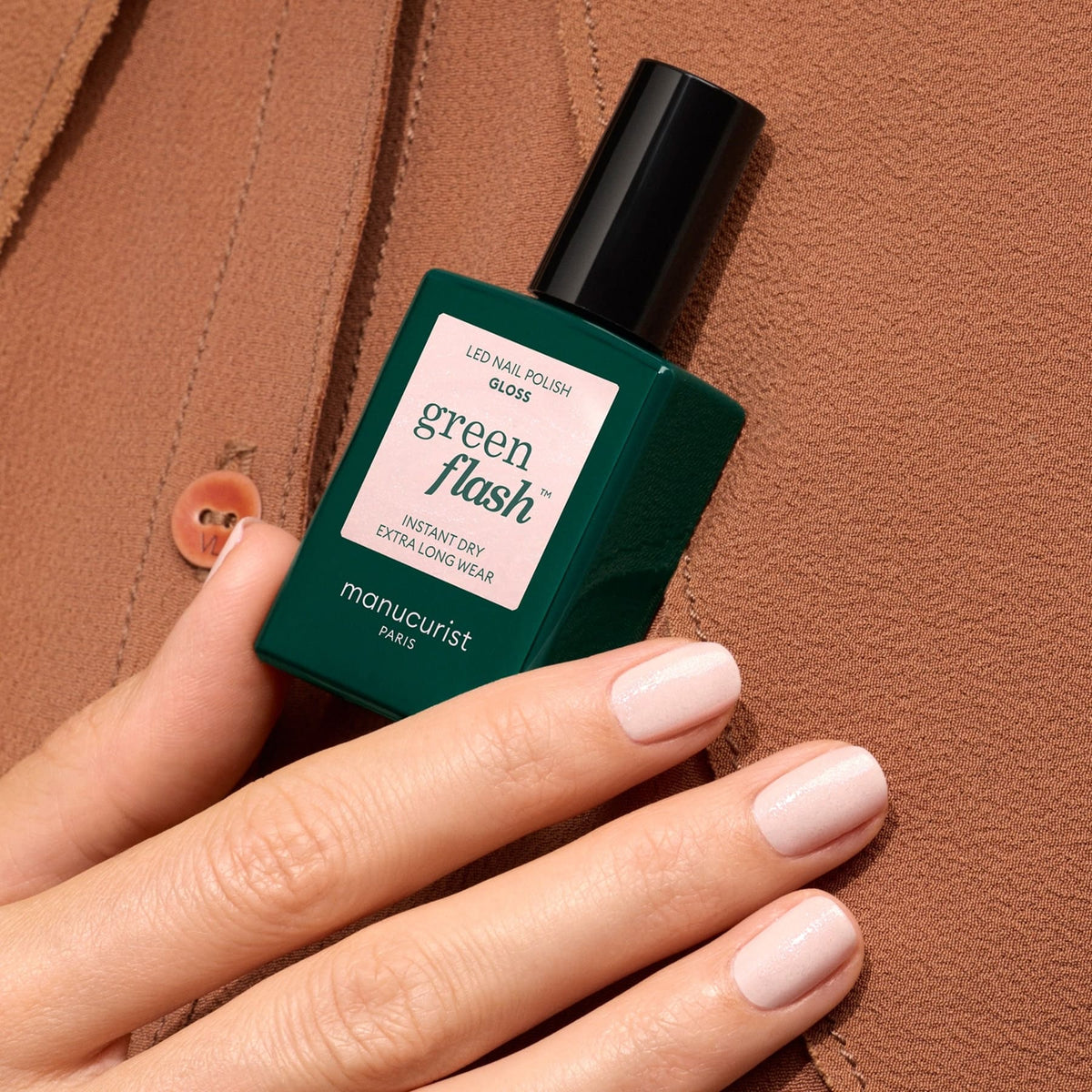 A hand with pale pink, iridescent-shimmer polished nails holds a green Manucurist Gloss bottle against textured brown fabric.