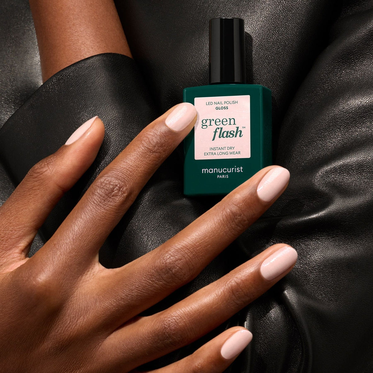 A hand with neatly manicured, iridescent pink shimmer nails rests on black leather next to a bottle of Manucurist Gloss.