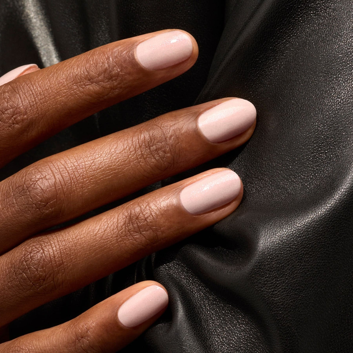 A hand with neatly manicured nails painted in Manucurist’s Gloss, an iridescent pink shimmer, rests on black textured fabric.
