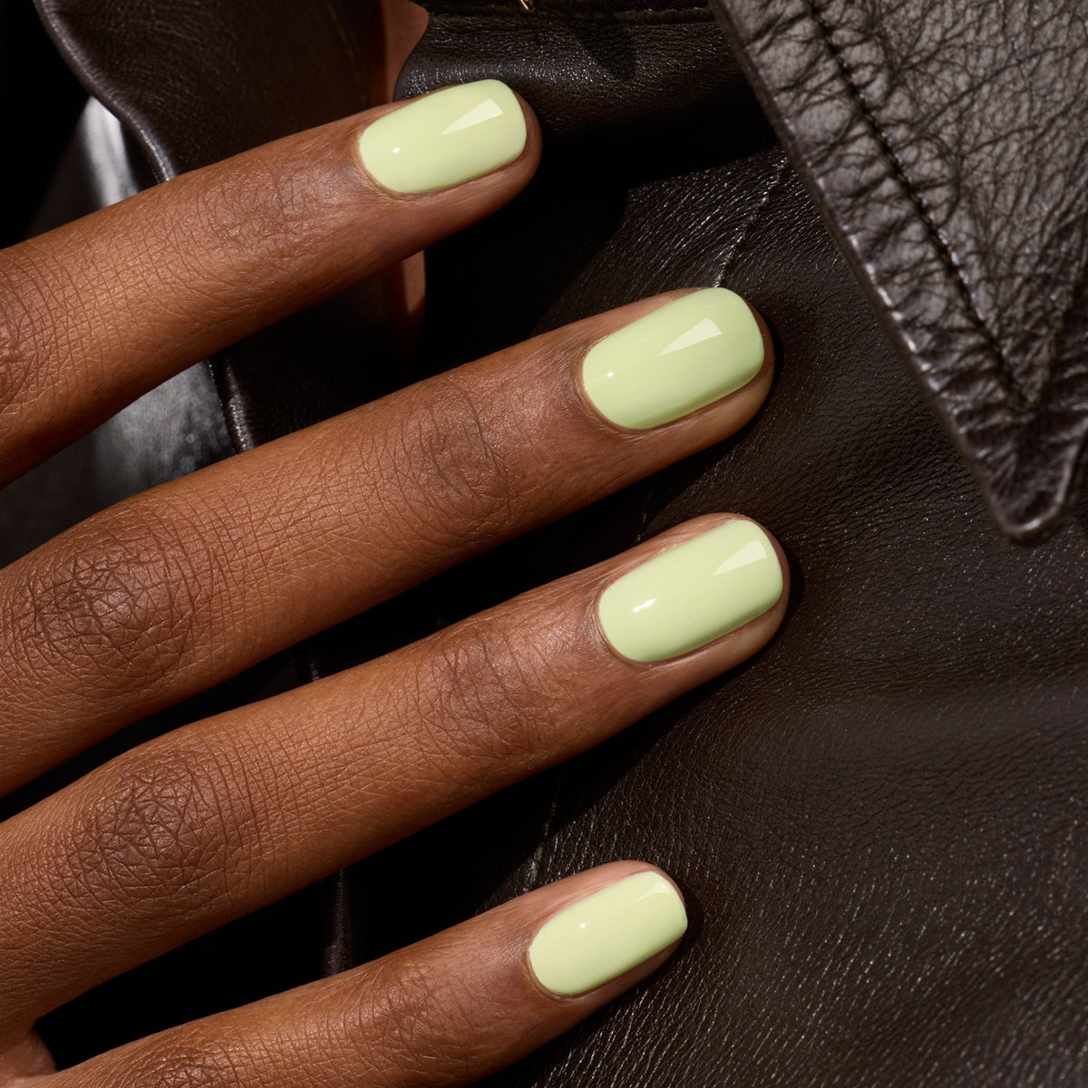 A hand with neatly manicured nails painted in Manucurist Pistachio, a pale green polish with subtle yellow undertones, rests on a black leather jacket.