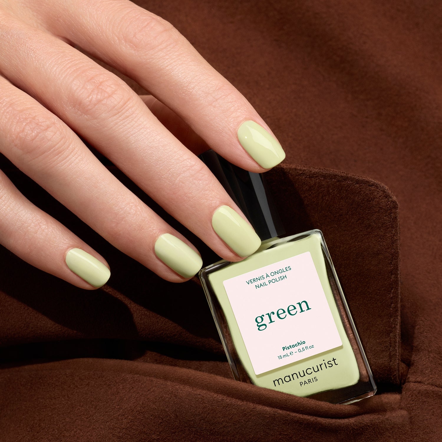 A hand with neatly manicured, pale green nails holds a matching bottle of Manucurist Pistachio nail polish against a soft brown fabric background.