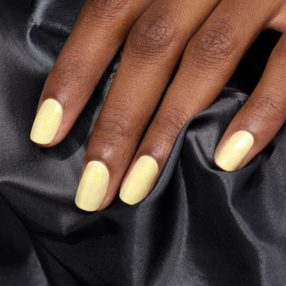 A hand with neatly manicured nails painted in Manucurist’s Lemonade, a pale yellow gel polish, rests on black satin fabric and showcases subtle glittering highlights.