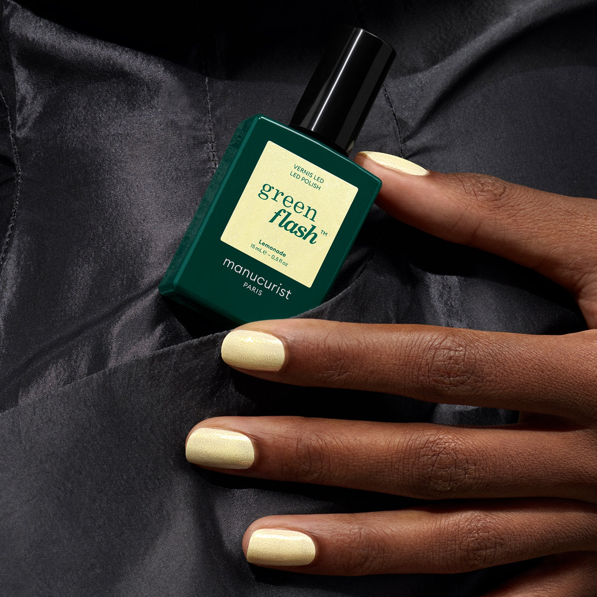 A hand with pale yellow gel polish holds a green bottle of Manucurist Lemonade nail polish against a dark, silky fabric background.