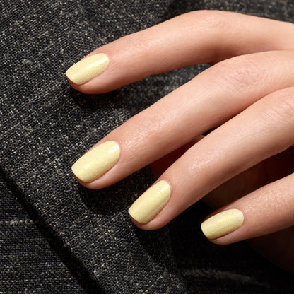 A close-up of a hand with neatly manicured nails painted in Manucurist’s Lemonade gel polish, a pale yellow shade with glittering highlights, resting on dark textured fabric.