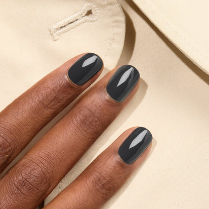 A close-up of three fingers wearing Manucurist’s Anthracite, a glossy dark grey shade from the Green™ range, resting on beige fabric with a buttonhole.