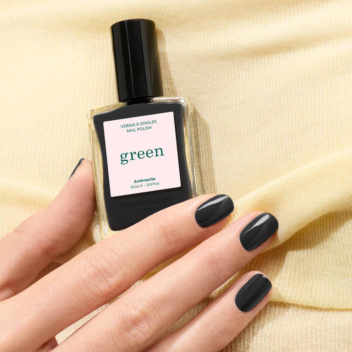A hand with short, glossy dark grey nails holds a bottle of Manucurist Anthracite nail polish against a soft yellow fabric background.