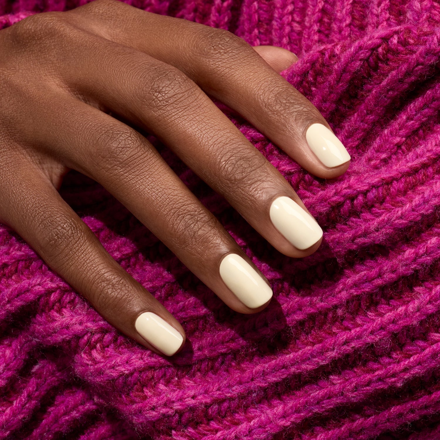 A hand with short, neatly manicured nails painted in glossy off-white Manucurist Shell LED gel polish rests on bright pink knitted fabric.