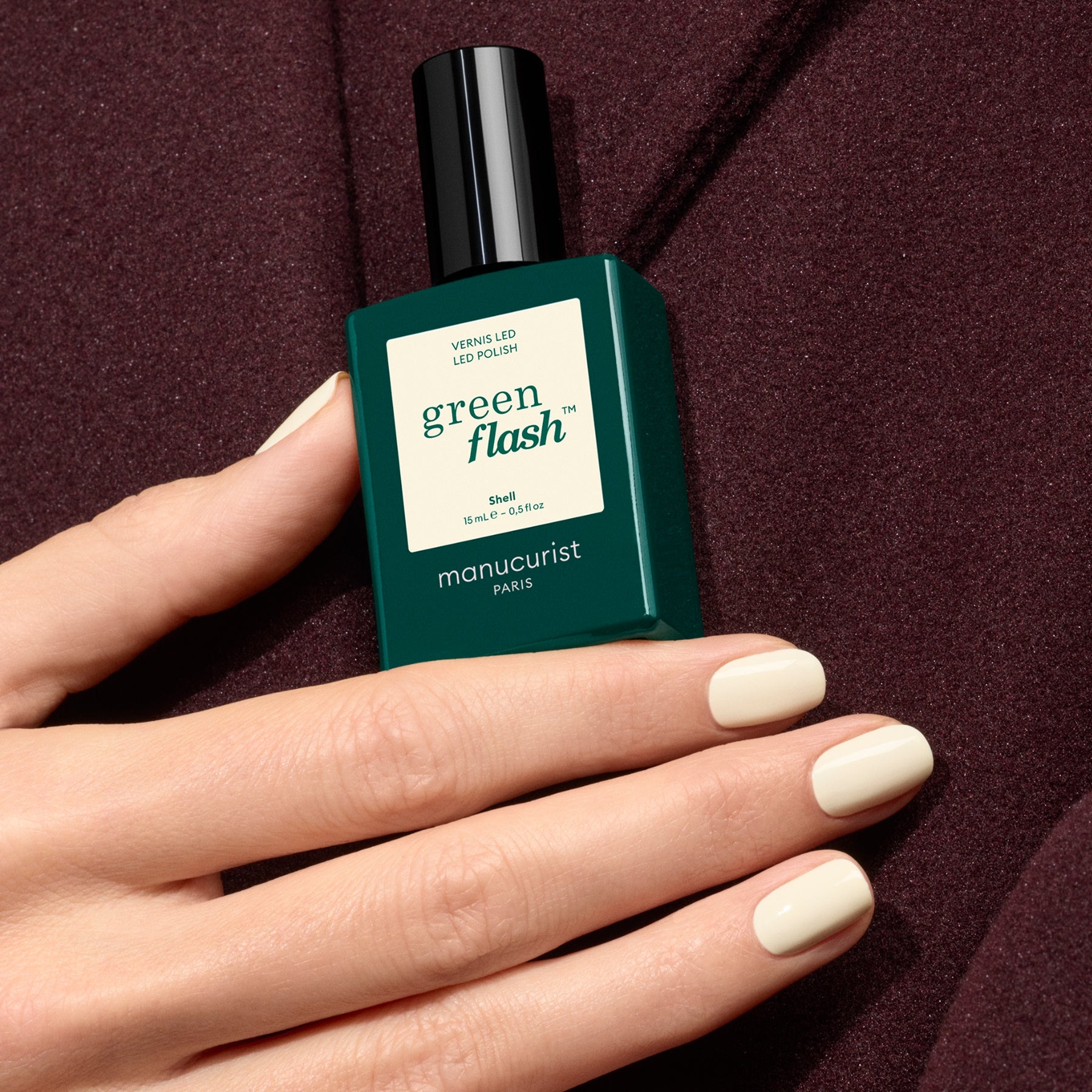 A minimalist hand with off-white nails holds a dark green bottle of Shell by Manucurist, set against burgundy fabric.