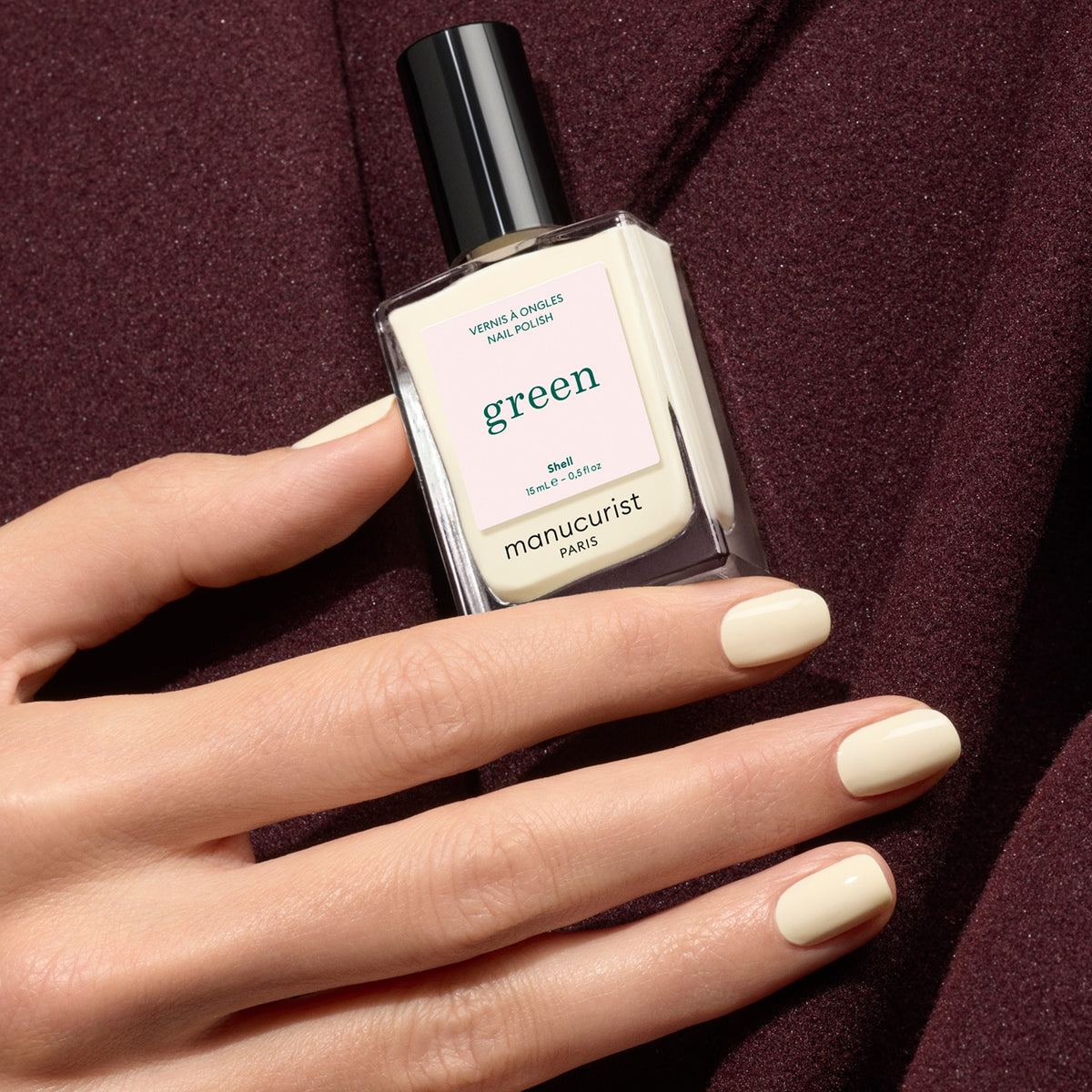 A hand with neatly manicured nails painted a light cream shade holds a bottle of Manucurist Shell nail polish against a burgundy fabric background.