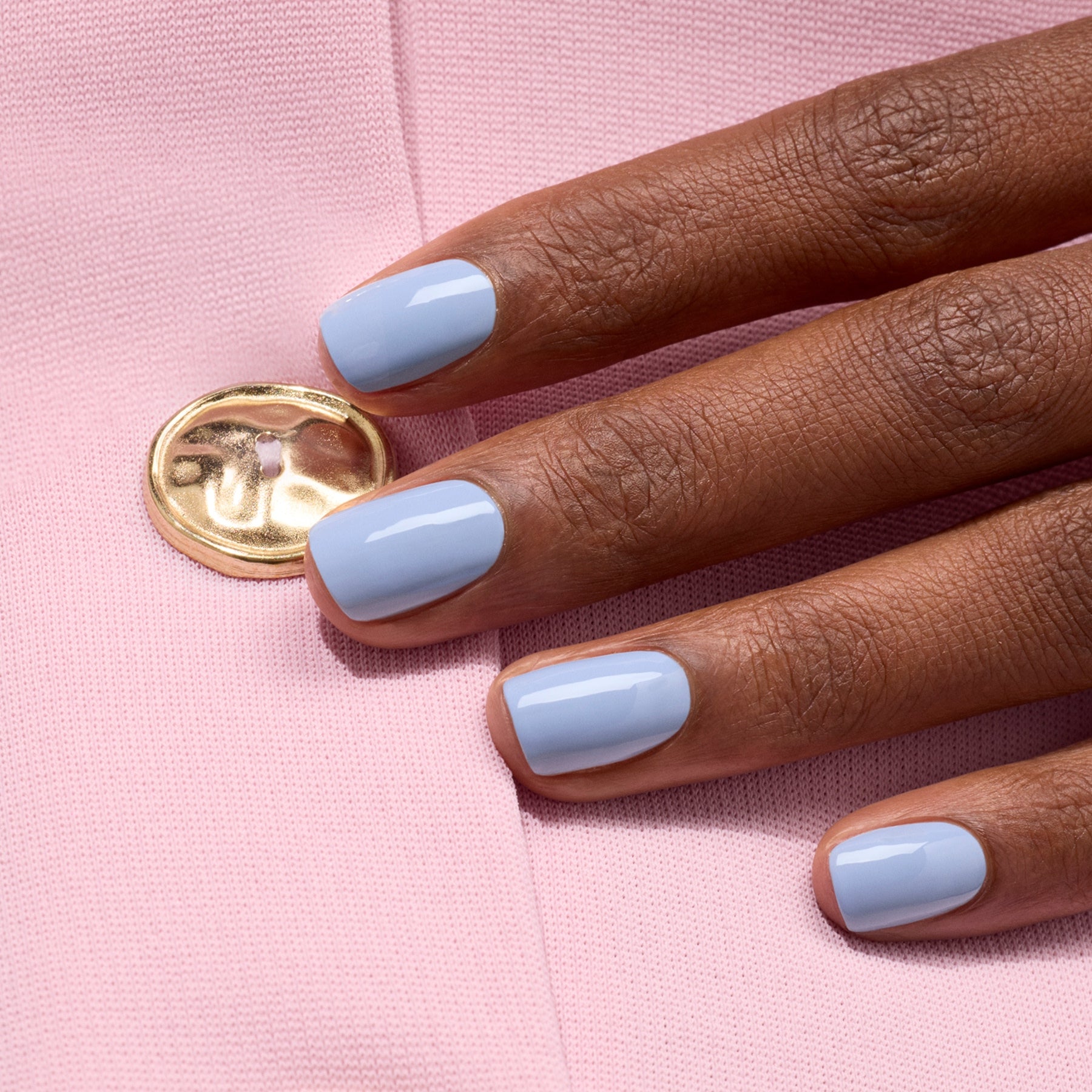 A hand with neatly manicured nails painted in glossy Manucurist Green Flash gel polish in the shade Lilas rests on light pink fabric with a shiny gold button.