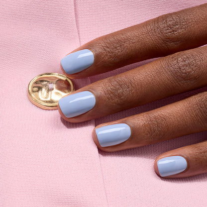 A hand with neatly manicured nails painted in glossy Manucurist Green Flash gel polish in the shade Lilas rests on light pink fabric with a shiny gold button.