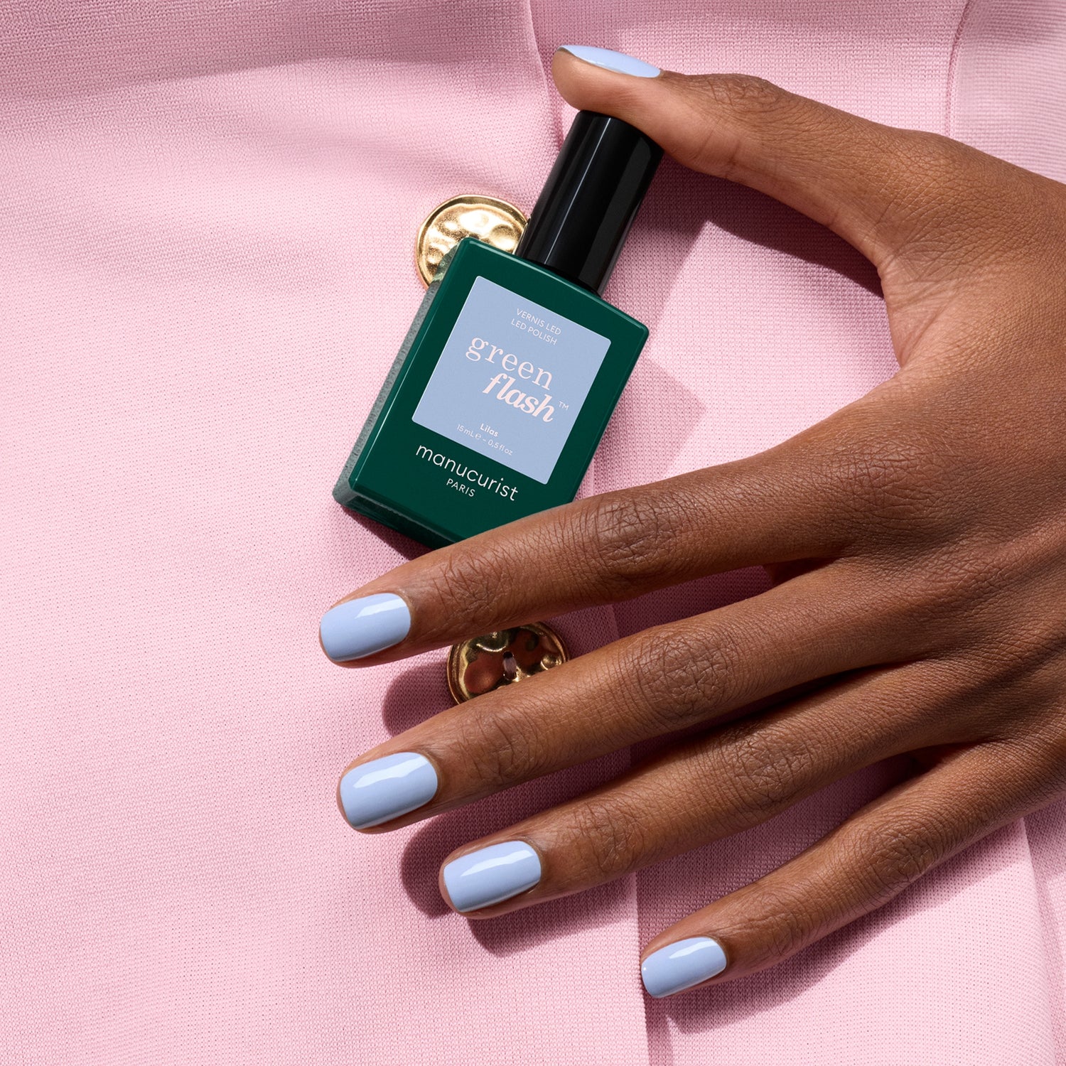 A hand with pastel blue gel polish holds a green bottle of Manucurist Lilas against a pale pink fabric background with a gold button.