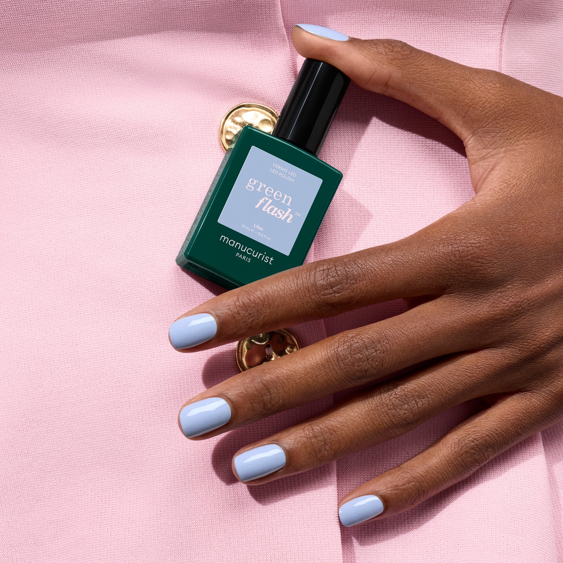 A hand with pastel blue gel polish holds a green bottle of Manucurist Lilas against a pale pink fabric background with a gold button.