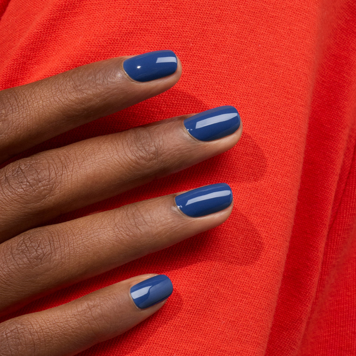 A hand with short, neatly manicured nails painted in glossy Midnight polish by Manucurist rests on a bright red fabric background.