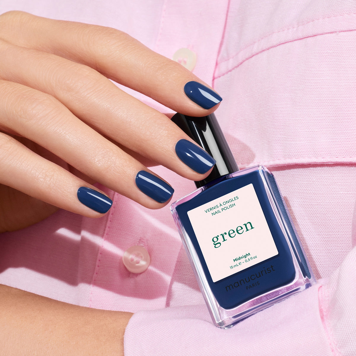 A hand with neatly manicured nails painted in Midnight by Manucurist holds a matching bottle of the polish against a light pink shirt.